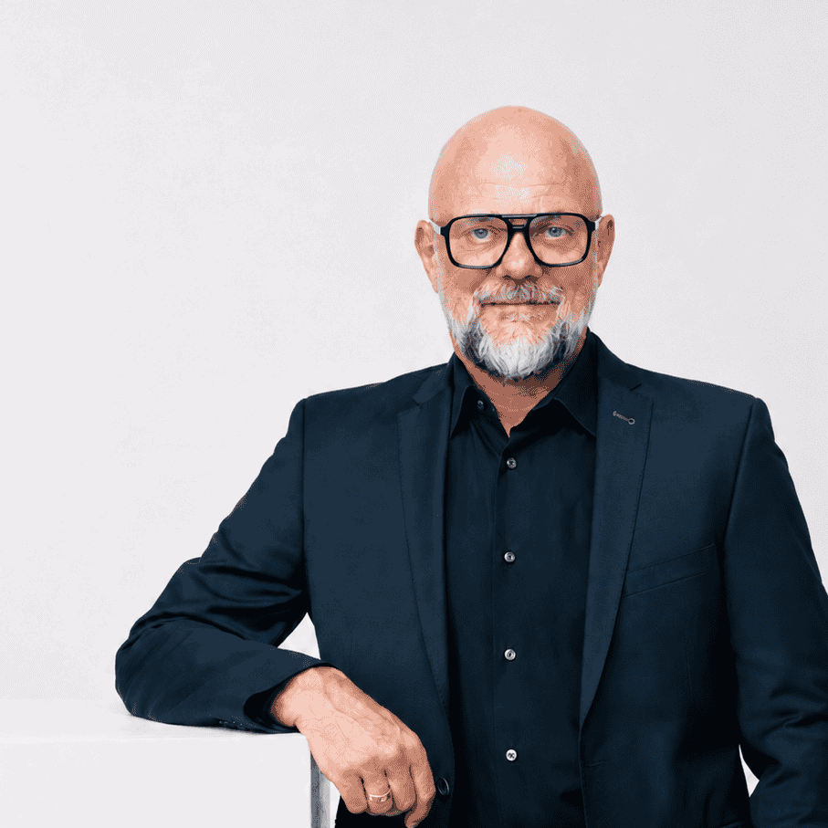 Älterer Mann in dunklem Anzug und Brille vor weißem Hintergrund.